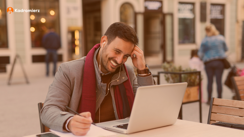 Mężczyzna siedzi przy stoliku na zewnątrz kawiarni. Pracuje przy laptopie, w ręku trzyma długopis. Uśmiecha się.