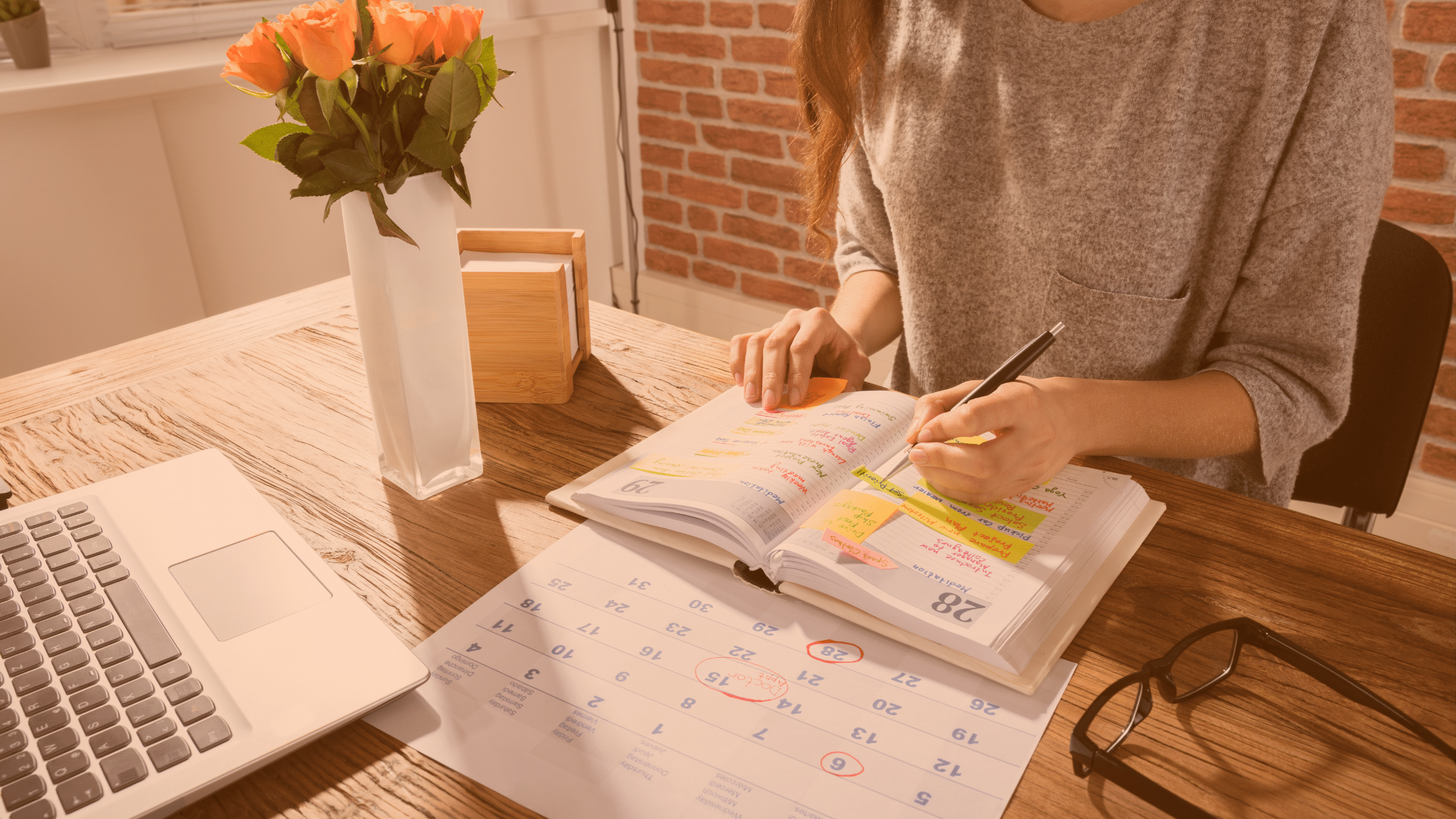 Przy biurku siedzi kobieta planująca harmonogram. Przed nią leży laptop oraz kalendarz miesięczny.