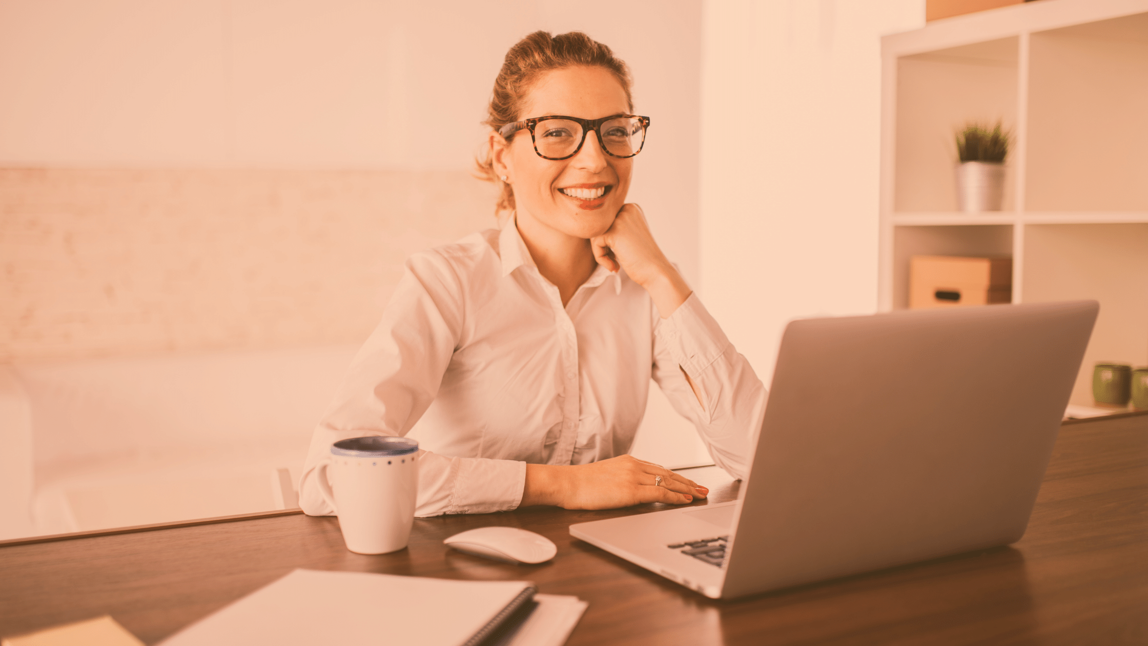 Kobieta siedzi przy biurku przy laptopie, uśmiecha się. Ma na sobie białą koszulę i okulary. 