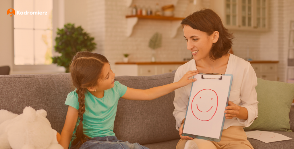 Dziewczynka wskazuje na rysunek uśmiechniętej postaci trzymany przez wychowawcę.
